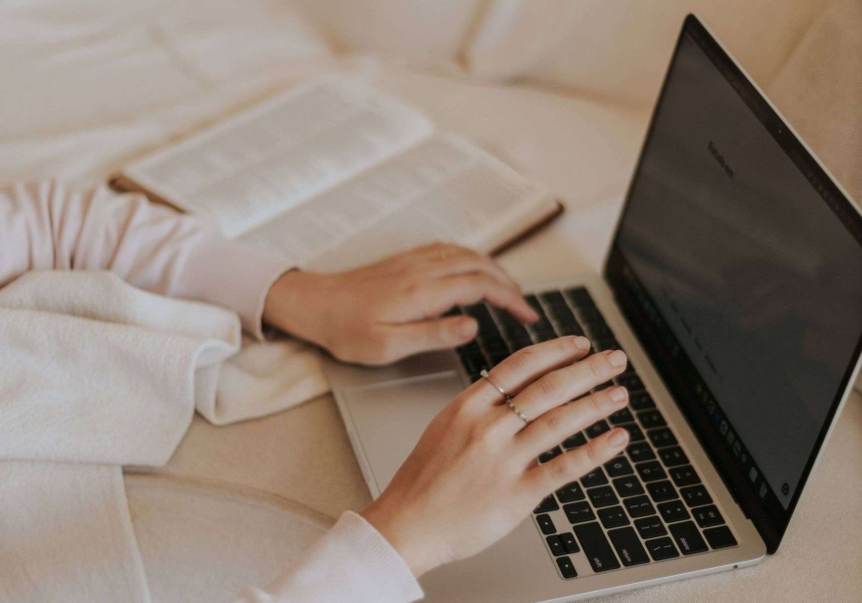 A cozy indoor setting with hands typing on a laptop, next to an open book on a soft surface.