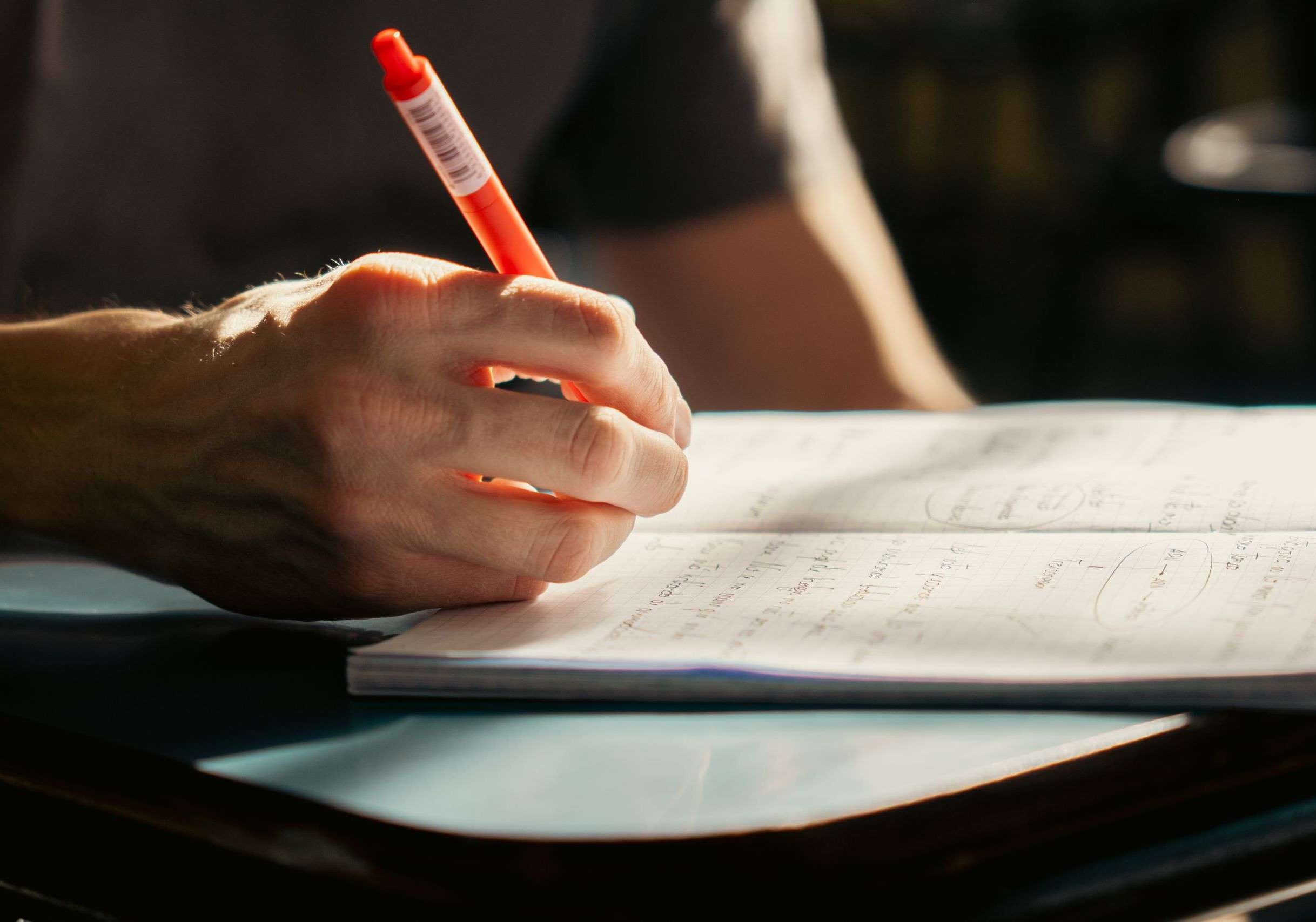 A student writing in a notebook with a pen, in a well-lit classroom environment.